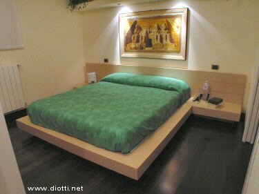 Il letto con comodini integrati Doge in rovere sbiancato. Molto raffinato l'abbinamento con il copriletto verde ed il quadro illuminato dai faretti nell'abbassamento.